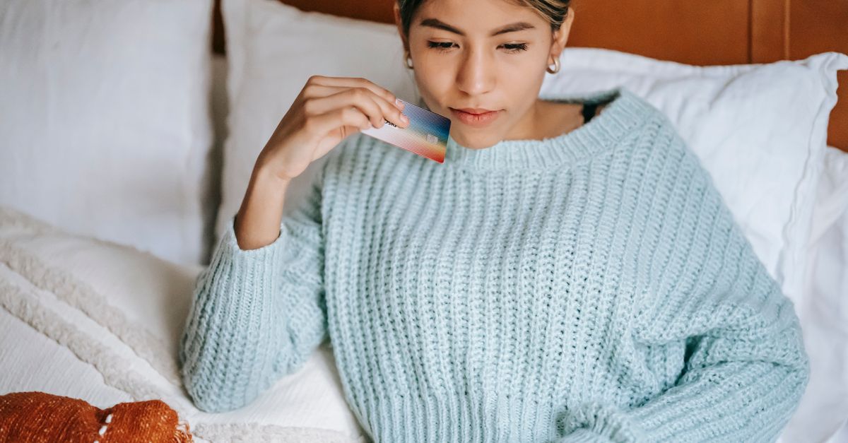 Crop concentrated Asian female in knitted blue sweater lying on cozy bed with credit card